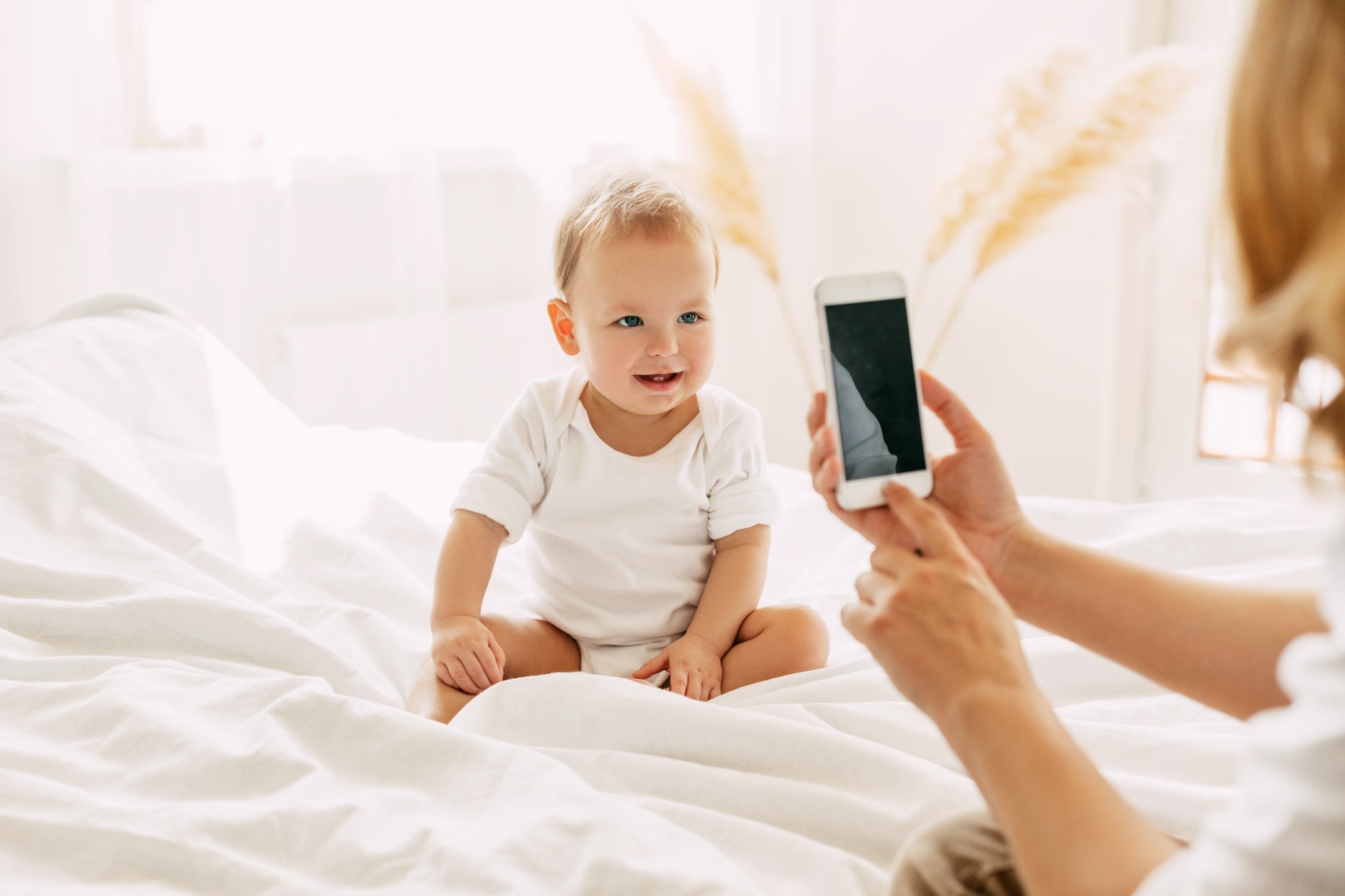 Cute baby is sitting on the bed and posing for his mom, mom is taking pictures of her baby